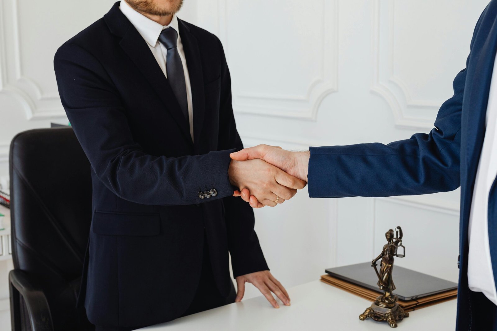 Nuestra Firma Two men in suits shaking hands in a formal office environment symbolizing an agreement.